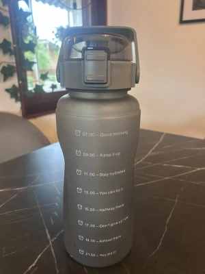 Grå vattenflaska med tidsmarkeringar - Snygg grå vattenflaska i plast med motiverande tidsmarkeringar och citat på framsidan. Flaskan har ett praktiskt lock med lås och bärhandtag, perfekt för att hålla koll på vätskeintaget under dagen.