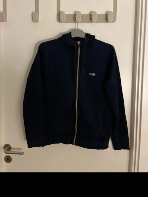 Hugo boss zip hoodie - Snygg mörkblå hoodie från Hugo Boss med dragkedja framtill och gul logga på bröstet. Hoodien har huva med dragsko och två fickor på sidorna. Tillverkad i mjukt bomullsmaterial som är skönt att ha på sig. Perfekt för en avslappnad och stilren look.