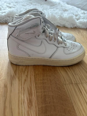 Nike Air Force 1 vita high tops - Klassiska vita Nike Air Force 1 high tops med snörning och kardborreband runt ankeln. 