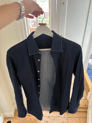 Mörkblå skjorta från Jacob Cohen - Snygg mörkblå denim skjorta från Jacob Cohen med diskreta ränder och klassisk krage. Skjortan har vita knappar och lång ärm, perfekt för en stilren look. Storlek 38 vilket motsvarar en S eller Slim M. Modellen på bilden är runt 190cm och väger 70kg. Nästan helt oanvänd, inga defekter. 