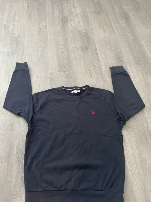 Mörkblå sweatshirt från U.S. Polo Assn. - Mörkblå sweatshirt från U.S. Polo Assn med rund halsringning och röd broderad logga på bröstet. Tröjan har långa ärmar, ribbade muddar och är gjord i mjukt bomullsmaterial. Enkel och stilren design som funkar till många outfits.