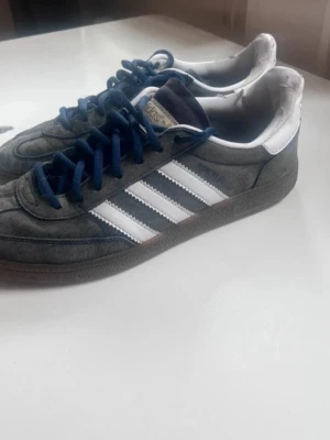 Adidas grå sneakers med blå detaljer - Säljer ett par klassiska Adidas sneakers i grå mocka med vita ränder på sidorna och blå skosnören. Skorna har låg profil och vit hälkappa. Perfekta för dig som gillar retrostil och streetwear.
