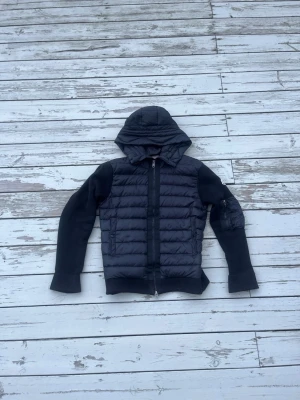 Svart Moncler cardigan - Snygg svart pufferjacka med huva och dragkedja framtill. Jackan har quiltade partier på framsidan och mjuka stickade ärmar och rygg. Perfekt för kyliga dagar när du vill ha både stil och komfort.