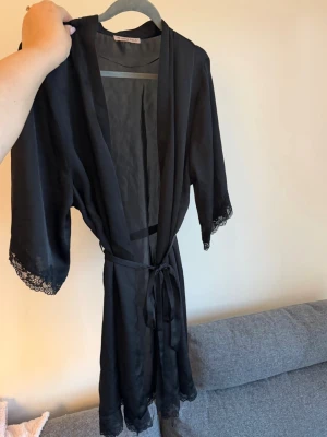 Svart kimono morgonrock med spets - Snygg svart kimono morgonrock i satinliknande material. Med fina spetsdetaljer vid ärmslut och nederkant. Knytband i midjan för justerbar passform. 