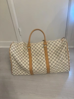 Rutigt weekendbag i beige och vitt - Snygg weekendbag med klassiskt rutmönster i beige och vitt, detaljer i ljusbrunt läder och två robusta handtag. Perfekt för resor och har en rymlig form. Väskan har dragkedja upptill och stilren design.