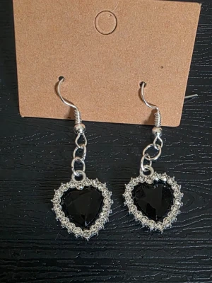 Handgjorda Hjärtformade örhängen med svarta stenar - Snygga handgjorda örhängen i silverfärg med hjärtformade svarta stenar i mitten, omgivna av små glittrande pärlor. Hängande modell med kedjedetalj och krokfäste. Perfekta för att ge din look en edgy touch.