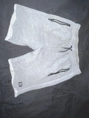Grå mjukisshorts med dragkedjor - Snygga grå mjukisshorts med resår i midjan och snörning. Shortsen har två fickor fram med svarta dragkedjor och en bakficka. Perfekta för chill eller träning. Tillverkade i mjukt bomullsmaterial.