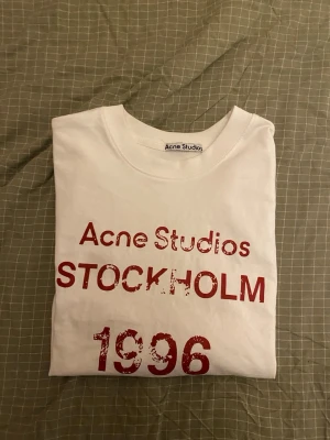 Acne studio t shirt - Vit t-shirt från Acne Studios med rött tryck på bröstet där det står 'Acne Studios STOCKHOLM 1996'. Klassisk passform och rund halsringning. Perfekt för dig som gillar enkel men snygg design och vill ha något från ett ikoniskt svenskt märke.