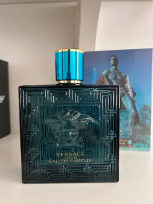 Versace Eros Eau de Parfum 100ml - Versace Eros Eau de Parfum i en lyxig, fyrkantig flaska i mörk turkos glas med Medusa-huvudet i relief på framsidan. Flaskan har ett elegant blått och guldigt lock och klassisk Versace-design. Doften är intensiv och maskulin, perfekt för dig som vill sticka ut.