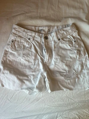 Vita högmidjade jeansshorts - Säljer ett par vita högmidjade jeansshorts från Pull&Bear. Shortsen har slitna detaljer framtill, fem fickor och knappgylf. Perfekta till sommaren och lätta att matcha med olika toppar.