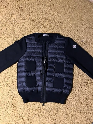 Svart dam-cardiganjacka från Moncler - Snygg svart cardiganjacka från Moncler med quiltad front och stickade ärmar. Jackan har två framfickor, dragkedja och Moncler-logga på ärmen. Materialmix med vadderad front och ribbade muddar ger en modern look. kvittot finns!!