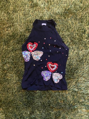 Svart paljettopp med hjärtan - Svart ärmlös topp med hög hals och stora paljettdetaljer i form av hjärtan och blommor framtill. Färgglada paljetter och broderade hjärtan i rött, lila och silver ger en unik look. Perfekt för fester, klubbar och om du vill se extra strålande ut!
