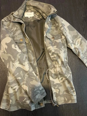 Camouflage vindjacka från Vila - Snygg vindjacka från Vila i grått camouflagemönster. Jackan har dragkedja, tryckknappar, två bröstfickor med lock och snörning i midjan för en schysst passform. Tillverkad i ett lätt och slitstarkt material, perfekt för blåsiga dagar.