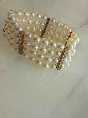 Brett pärlarmband med strassdetaljer - Snyggt brett armband med flera rader vita pärlor och guldfärgade detaljer med infällda strass. Armbandet har en elastisk design som gör det enkelt att ta på och av. Perfekt för dig som vill ha ett statement-smycke med lyxig känsla.
