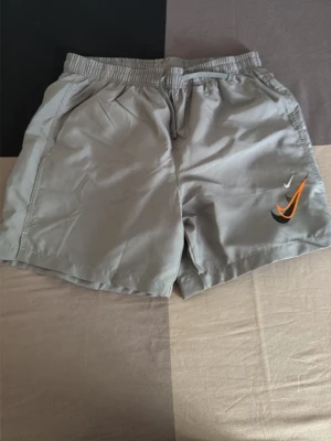 Grå badshorts från Nike - Säljer ett par grå badshorts från Nike med elastisk midja och snörning. Shortsen har fickor på sidorna och en bakficka med knapp. Nike-loggan finns framtill med orange och svart detalj. Perfekta för stranden eller poolen.