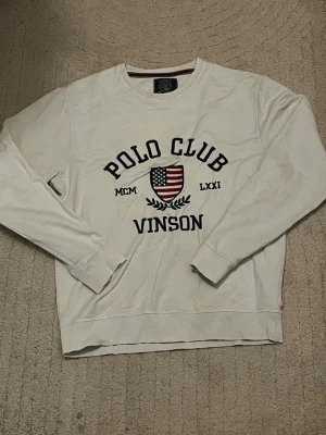 Vit sweatshirt från Vinson Polo Club - Vit sweatshirt med rund hals från Vinson Polo Club. Tröjan har svart text och ett broderat emblem med amerikanska flaggan på bröstet. Långa ärmar och ribbade muddar vid ärmslut och nederkant. Perfekt för en sportig och avslappnad stil.