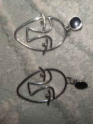 Silvriga ansiktsörhängen i metall - Unika örhängen i silverfärgad metall med abstrakt ansiktsdesign. Örhängena har en öppen, konstnärlig form och små svarta detaljer upptill. Perfekta för dig som gillar statement-smycken och modern stil.