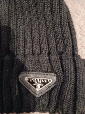 Svart ribbstickad mössa från Prada - Cool svart ribbstickad mössa från Prada med en stor fluffig svart pompom på toppen och ikonisk metalltriangel-logga framtill. Mössan har uppvikt kant och är tillverkad i mjukt stickat material som håller dig varm och snygg under kalla dagar.