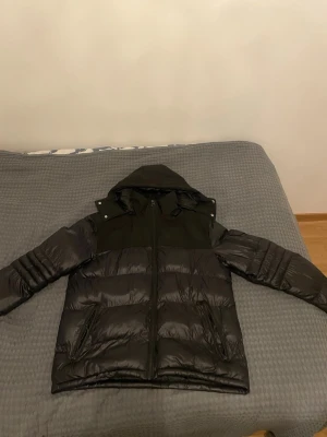 Svart pufferjacka med huva - Snygg svart pufferjacka med huva och dragkedja framtill. Jackan har quiltad design och två fickor på sidorna. Perfekt för kalla dagar och har en modern look som passar till det mesta.