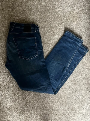 Mörkblå jeans från Replay - Snygga mörkblå jeans från Replay med klassisk femficksdesign och diskreta slitningar framtill. Jeansen har raka ben och är tillverkade i ett mjukt denimtyg som ger en skön passform. Perfekta för en avslappnad och trendig look.