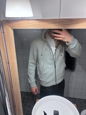 Grå zip hoodie från Burberry - Snygg grå zip hoodie från Burberry med klassisk huva och dragsko. Hoodien har två stora fickor fram och diskret Burberry-logga på bröstet. Tillverkad i mjukt bomullsmaterial och har ribbade muddar vid ärmslut och nederkant.