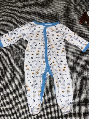 Vit sparkdräkt med blå detaljer - Söt vit sparkdräkt med blå kant och tryckknappar framtill. Mönstrad med text som 'LOVE', 'OMG', små bin, tassavtryck, kronor och blommor i blått och gult. Långa ärmar och täckta fötter. Perfekt för mysiga dagar hemma.