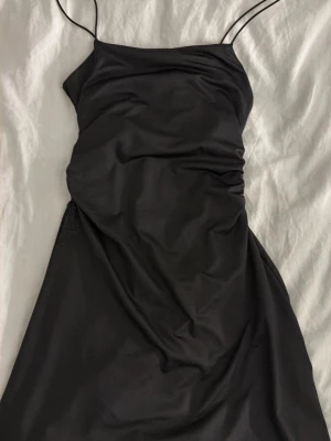 Svart kort klänning med tunna band - Stilren svart kort klänning med tunna axelband och figurnära passform. Klänningen har en enkel design utan mönster och är tillverkad i ett mjukt, slätt material som ger en elegant look.