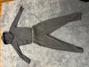 Grå mjukisdress med huva - Mysig grå mjukisdress i två delar med dragkedja framtill och huva. Byxorna har resår i midjan och muddar vid bensluten. Perfekt för chill hemma eller när du vill vara bekväm. Materialet känns mjukt och stretchigt.