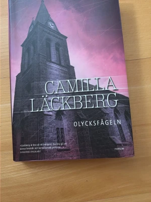 Olycksfågeln av Camilla Läckberg - Säljer boken 'Olycksfågeln' av Camilla Läckberg. Hård pärm med ett omslag i mörka toner och lila nyanser, motiv av en kyrka mot dramatisk himmel. Perfekt för dig som gillar spännande deckare och svensk krim.