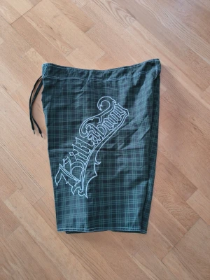 Rutiga shorts från Billabong - Snygga shorts från Billabong i svart och mörkgrönt rutigt mönster med stort vitt logotryck på sidan. Klassisk modell med dragsko i midjan och längd till knät. 