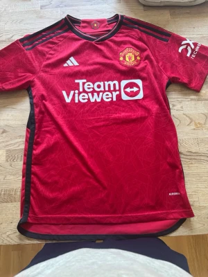 Manchester United matchtröja röd Adidas - Säljer en röd Manchester United matchtröja från Adidas med svarta detaljer och klubbmärke på bröstet. Tröjan har TeamViewer som sponsortryck, klassiska Adidas-ränder på axlarna och är tillverkad i lätt funktionsmaterial. Perfekt för fotbollsträning eller supporterliv.