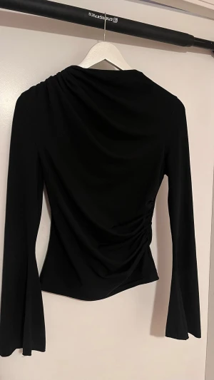 Svart långärmad topp med drapering - Snygg svart långärmad topp med draperad detalj på ena sidan som ger en cool twist. Figurnära passform och lätt utsvängda ärmar. Perfekt att styla med jeans eller kjol för en trendig look.