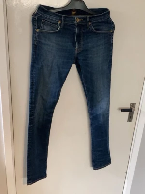 Blå raka jeans från Lee - Snygga blå jeans från Lee med klassisk femficksmodell och raka ben. Jeansen har guldfärgade detaljer vid fickorna och en brun läderpatch med Lee-loggan bak i midjan. Tillverkade i slitstarkt denimtyg som ger en tidlös look.