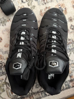 Nike Air Max Plus Tn Svart - Säljer ett par svarta Nike Air Max Plus Tn sneakers med ikoniska Tn-detaljer på hälen och reflekterande inslag på ovandelen. Skorna har en sportig siluett, mesh och syntetmaterial samt tydliga Air Max-dämpningar i sulan. Perfekta för dig som gillar streetwear.
