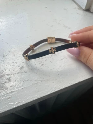 Svart läderarmband Tory Burch - Snyggt svart och brunt armband i läder från Tory Burch med guldfärgade logodetaljer runt om och ett matchande guldfärgat spänne. Perfekt accessoar för dig som gillar stilrena och exklusiva smycken med en twist.