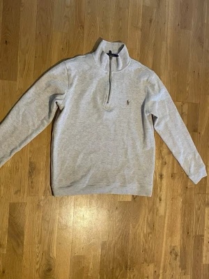 Ljusgrå half zip tröja från Polo Ralph Lauren - Ljusgrå långärmad tröja från Polo Ralph Lauren med klassisk half zip och liten broderad logga på bröstet. Tröjan har hög krage och är tillverkad i mjuk bomull, perfekt för en clean och avslappnad stil.