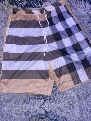 Beige och vita rutiga shorts - Säljer ett par snygga shorts med beige, vita och bruna rutor. Shortsen har resårmidja med vit dragsko och sidofickor. Perfekta för sommaren och riktigt sköna att bära till vardags. Materialet känns lätt och luftigt.