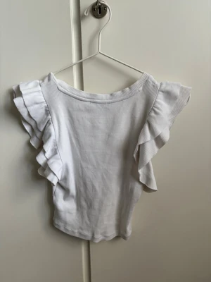 Vit ribbad topp med volangärmar - Supersöt vit ribbad topp med volangdetaljer på ärmarna. Toppen har en rund halsringning och är tillverkad i ett mjukt, stretchigt material som känns bekvämt mot huden. Perfekt för dig som gillar en feminin och stilren look.