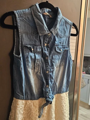 Ärmlös jeansklänning med spetskjol - Snygg ärmlös klänning med överdel i blått jeansmaterial och knappar framtill. Två bröstfickor med lock och en knytning nedtill. Kjolen är vit med spetsdetaljer som ger en romantisk touch. Perfekt för dig som gillar en mix av denim och spets.