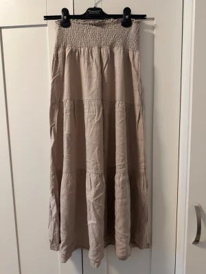 Beige linnekjol med smock i midjan - Luftig beige långkjol med smock i midjan och volangdetaljer. Kjolen har en avslappnad passform och är tillverkad i ett mjukt bomullsliknande material. Perfekt för varma dagar och enkel att styla med olika toppar.