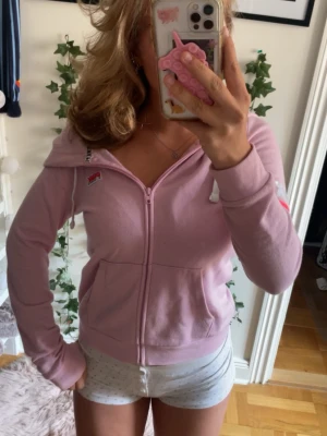 Rosa zip-hoodie från PINK - Säljer en ljusrosa hoodie från PINK med dragkedja framtill, vita snören i huvan och fickor på sidorna. Detaljer med logga på bröstet och en rosa/vit rand på ärmen. Perfekt för en chill och avslappnad stil.
