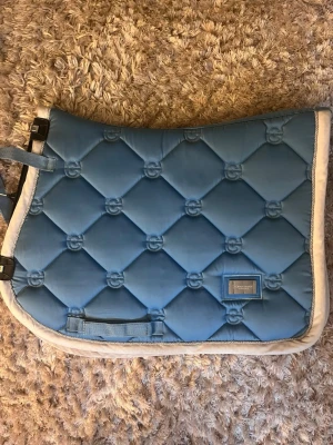 Blått schabrak från Equestrian Stockholm - Snyggt ljusblått schabrak från Equestrian Stockholm med quiltat mönster och vita kanter. Tillverkat i glansigt material med broderade detaljer och metallbricka med logga. Perfekt till hästutrustningen för dig som gillar stilrena accessoarer.