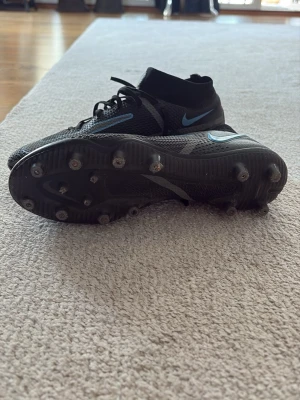 Nike Phantom svart/blå fotbollsskor - Säljer ett par Nike Phantom fotbollsskor i svart med blå detaljer och Nike-logga. Skorna har snörning, högre strumpkrage och fasta dobbar för bra grepp på planen. Tillverkade i syntetmaterial för lätt känsla och bra bollkontroll.