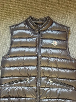 Svart dunväst från Moncler - Snygg svart dunväst från Moncler med glansig finish och klassisk logotyp på bröstet. Västen har ståkrage, två dragkedjefickor och quiltad design. Perfekt för lager-på-lager och streetstyle.
