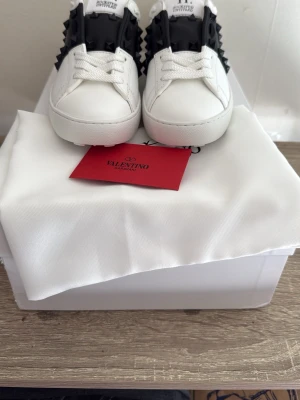 Valentino vita sneakers med nitar - Snygga vita sneakers från Valentino med svart detalj och coola nitar på sidorna. Skorna är i läder och har klassisk låg modell med vita snören och diskret logga på plösen. Perfekta för dig som vill sticka ut med en lyxig och edgy stil.