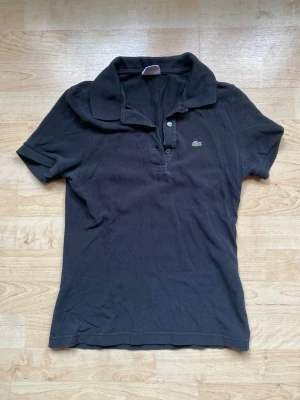 Svart polotopp från Lacoste - Klassisk svart polotopp från Lacoste med korta ärmar och krage. Toppens material är bomull och den har knappar framtill samt den ikoniska Lacoste-loggan på bröstet. Perfekt för en clean och sportig look.
