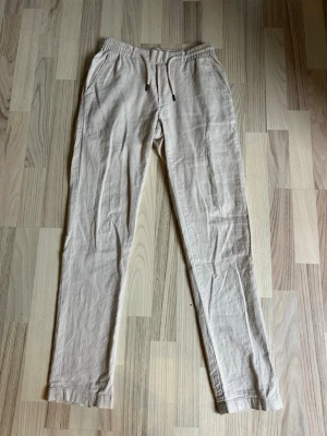 Beiga linnebyxor - Snygga beiga linnebyxor med avslappnad passform och elastisk midja. Byxorna har sidfickor fram och två bakfickor med knapp. Perfekta för en chill och stilren look. Tillverkade i luftigt linnematerial.