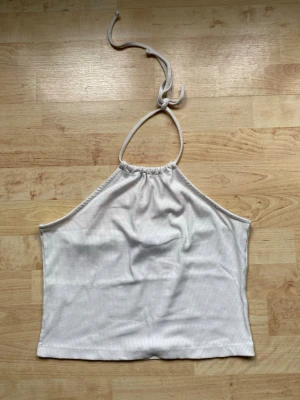 Vit ribbad halterneck linnetopp - Säljer en vit ribbad linnetopp med halterneck och knyt i nacken. Toppen är ärmlös och har en croppad passform som passar perfekt till sommaren. Enkel och stilren design utan synliga loggor eller mönster.