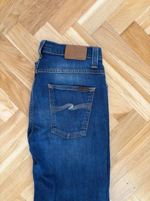 Blå jeans från Nudie Jeans - Säljer ett par klassiska blå jeans från Nudie Jeans med raka ben och snygga kontrastsömmar. Jeansen har två bakfickor med den ikoniska Nudie-bågen och patch i brunt läder bak i midjan. Tillverkade i slitstark denim, perfekta till vardagslooken.
