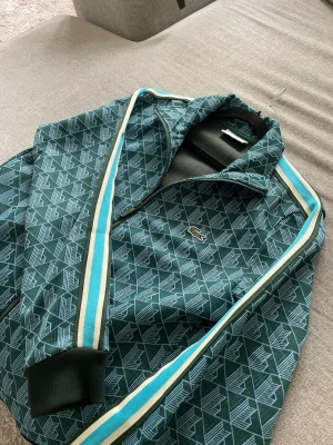 Grön mönstrad hoodie från Lacoste - Snygg grön hoodie från Lacoste med ett grafiskt mönster i vitt och grönt. Tröjan har dragkedja framtill, ribbade muddar och vita samt turkosa ränder längs ärmarna. Klassisk Lacoste-logga på bröstet. Perfekt för dig som gillar streetwear och sportig stil.